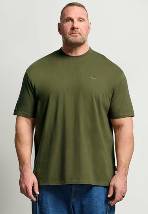 Voksen muskuløs mand med kort hår og skæg iført en olivengrøn T-shirt og blå jeans, stående mod en ensfarvet lys baggrund.