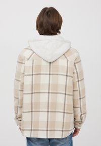 Giacca flanella a quadri in beige e crema con cappuccio grigio. Presenta una vestibilità rilassata, maniche lunghe e polsini con bottone per la regolazione.