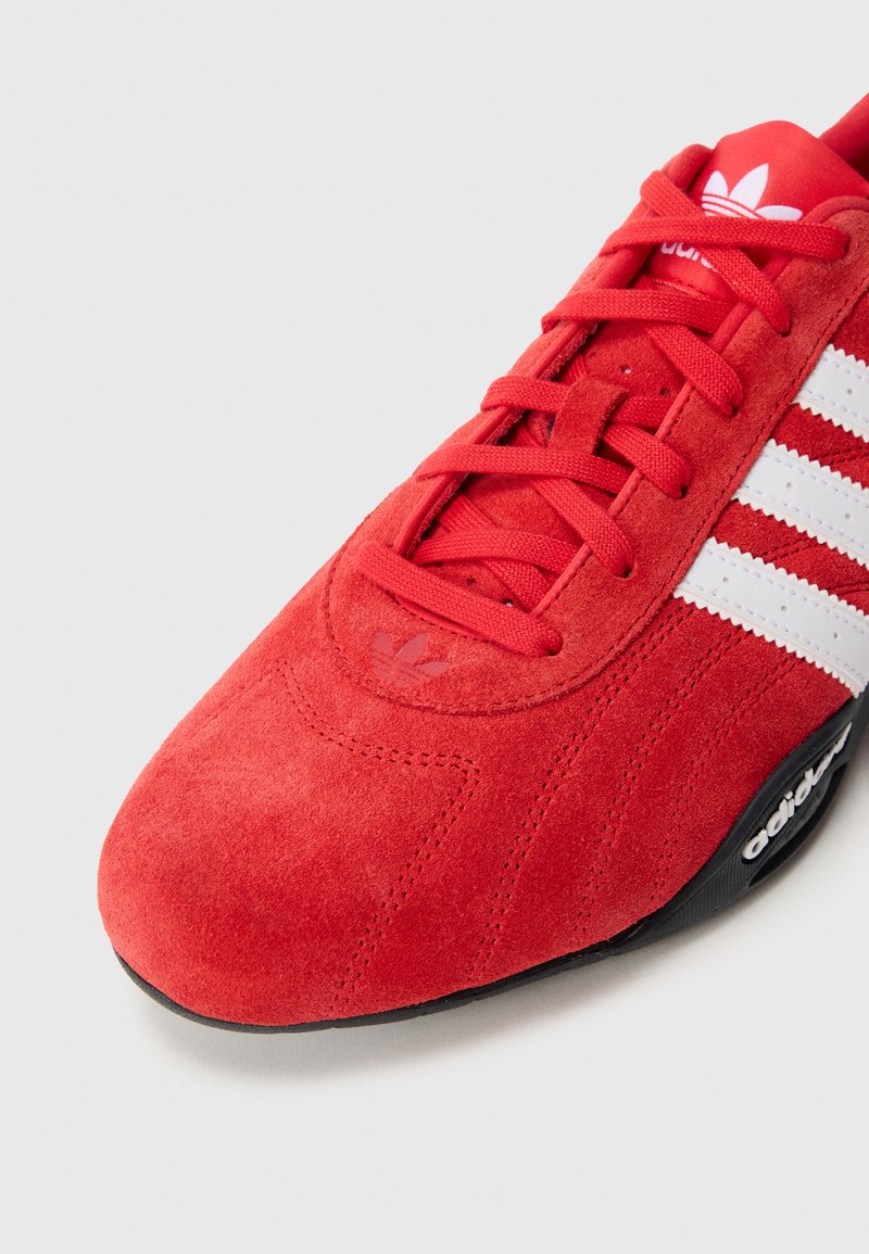 Baskets en daim rouge avec un design à trois bandes blanches, présentant des accents texturés et une semelle noire avec logo. Les lacets sont assortis à la couleur des chaussures.