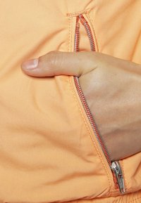 Tessuto di giacca arancione chiaro con una tasca con zip, zip a contrasto di un arancione più scuro, e una mano che afferra la zip per facilitarne l'accesso.