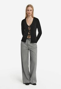 Svart långärmad cardigan med djup V-ringning och snörning fram. Tillsammans med grå vida byxor med ett subtilt randmönster.