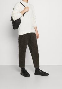 Krämfärgad stickad tröja, svarta manchesterbyxor och svarta sneakers. En svart ryggsäck hänger över ena axeln. Släta, strukturerade tygdetaljer.