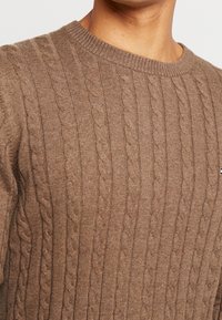Pull en tricot marron avec un col côtelé et un motif texturé, mettant en avant un tissu doux et confortable. Présente un accent de marque subtil.