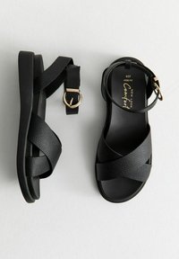 Las sandalias negras presentan un upper de cuero texturizado con tiras entrelazadas, una correa en el tobillo con una hebilla en tono dorado y una suela plana.