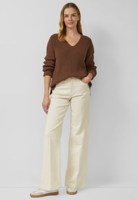 Frau steht, trägt einen braunen V-Ausschnitt-Strickpullover, cremefarbene Weitbein-Hosen und weiße Turnschuhe vor einem schlichten hellgrauen Hintergrund.