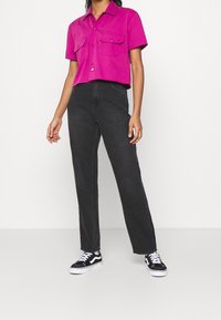 Camicia rosa a maniche corte con due tasche sul petto, abbinata a jeans neri a vita alta e sneakers nere con suola e lacci bianchi.