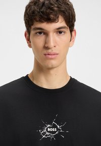 Fiatal férfi rövid, göndör barna hajjal, fekete pólót visel, amelynek közepén a "BOSS" felirat és egy fehér repedt üveg mintázat látható.