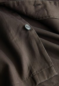 Gros plan de tissu brun foncé avec un bouton gris à quatre trous cousu près d'une boutonnière sur un bord de vêtement cousu.