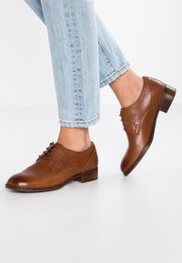 Chaussures oxford en cuir marron avec un design à lacets, petit talon empilé, détails perforés et texture lisse, assorties à un jean bleu clair.
