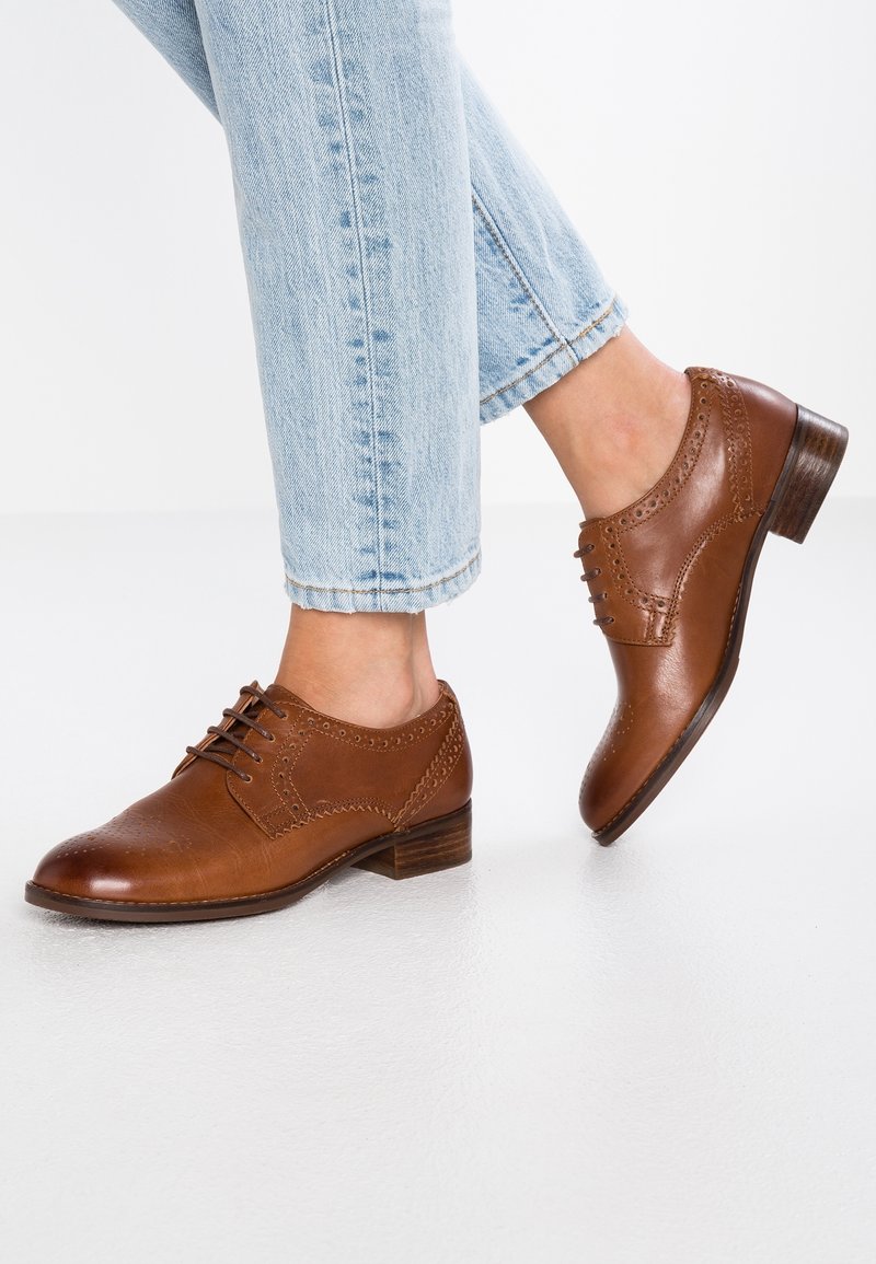 Chaussures oxford en cuir marron avec un design à lacets, petit talon empilé, détails perforés et texture lisse, assorties à un jean bleu clair.