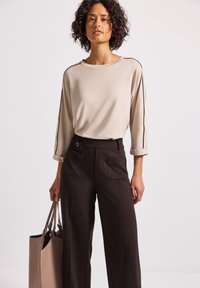 Haut beige à manches longues avec des rayures noires sur les côtés, associé à un pantalon large marron avec poches avant et un accent en forme d'anneau argenté ; portant un sac tote beige.