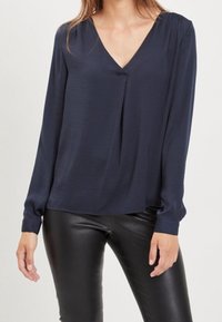 Mörkblå långärmad blus med V-ringning, som har en lös passform och subtil drapering framtill, kombinerad med svarta läderbyxor.