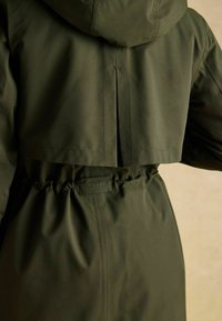 Zielona kurtka z kapturem, z wąską talią, żebrowanym materiałem oraz detalem w postaci plis na plecach. Zawiera widoczną metkę Joules na talii.