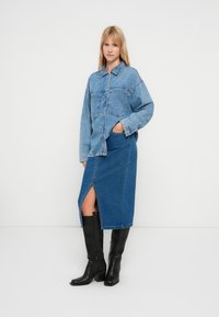 Light blue denim jacket with a front pocket, paired with a knee-length blue denim skirt featuring a slit, and black knee-high boots.