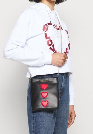 Personne portant un sweat à capuche blanc avec du texte rouge "LOVE MOSCHINO", un jean bleu et tenant un sac à bandoulière noir orné de trois patchs en forme de cœur rouge.