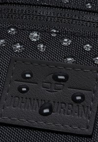 Černá texturovaná tkanina s kapkami vody na povrchu, s obdélníkovou černou koženou etiketou s vyraženým nápisem "JOHN URBAN."