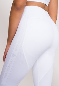 Leggings atletici bianchi con vita a coste, design testurizzato e marchio discreto lungo il lato. Il materiale sembra elastico.