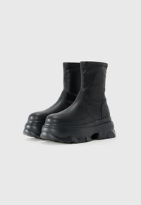 Botas negras de media caña con un suave cuero en la parte superior y suelas gruesas de plataforma con estrías, exhibidas sobre un fondo blanco liso.