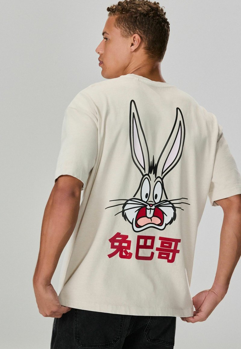 Cremfarbener T-Shirt mit einem Grafik von Bugs Bunny, der lange Ohren und ausdrucksstarke Merkmale hat, Akzentuiert mit rotem Text in chinesischen Zeichen.