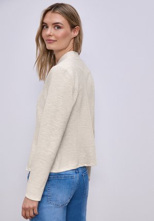 Jeune femme aux longs cheveux blonds, vêtue d'une veste crème texturée et d'un jean bleu, regardant par-dessus son épaule sur un fond uni.