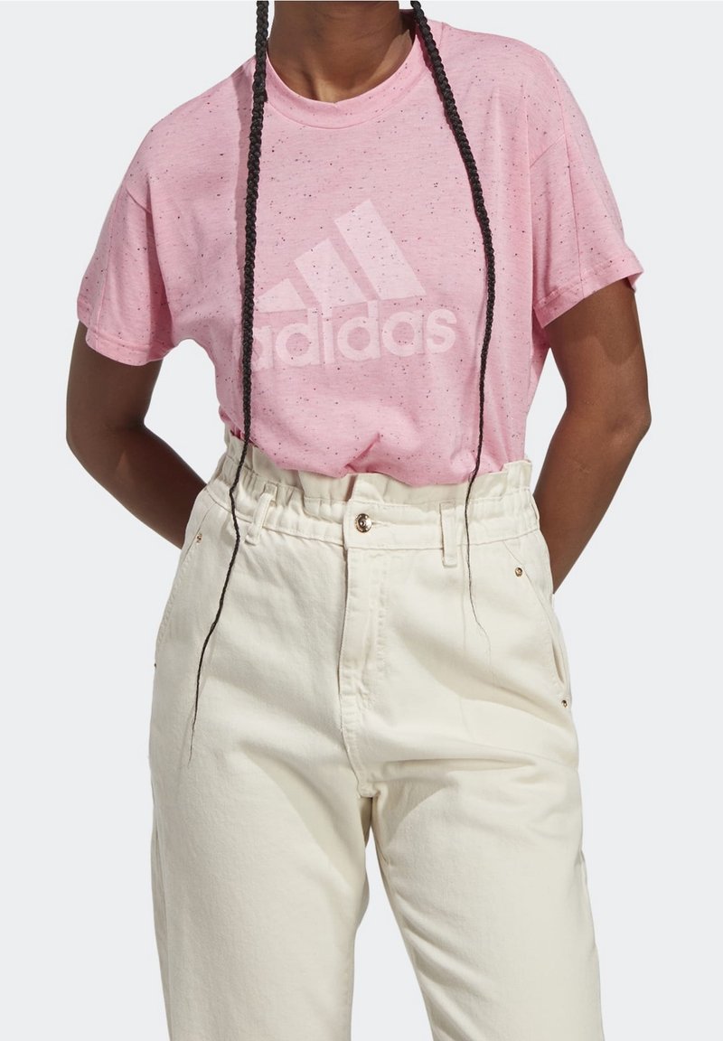 Vaaleanpunainen t-paita, jossa on pilkullinen rakenne ja suuri valkoinen Adidas-logo, yhdistetty korkeavyötäröisiin kermaisenvärisiin housuihin.