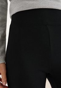 Sorte leggings lavet af strækbart stof, der har en glat tekstur og en høj talje. Overdelen er en lysegrå, langærmet skjorte.