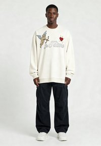 Homme portant un sweat blanc orné d'un chérubin, d'un cœur rouge et du texte "Je T'aime", un pantalon cargo noir et des baskets blanches, debout devant un fond uni.