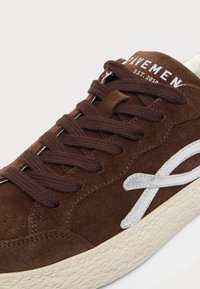 Baskets en daim marron avec lacets marron foncé, semelle blanche texturée et logo incurvé blanc brodé sur le côté.