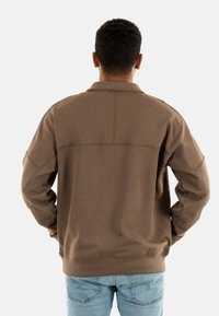 Felpa marrone con colletto alto, spalle drop e polsini a coste. Texture liscia e dettagli di cucitura minimi. Indossata con jeans azzurri chiari.
