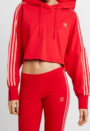 Kobieta ubrana w czerwoną krótką bluzę z kapturem oraz dopasowane legginsy z wysokim stanem, ozdobione różowymi paskami i logo Adidas na piersi oraz udzie.