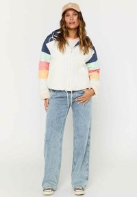 Polstret, hvid jakke med farverige stribede ærmer i pink, blå og grøn; parret med lyseblå jeans og sneakers. Justerbar snor i taljen.