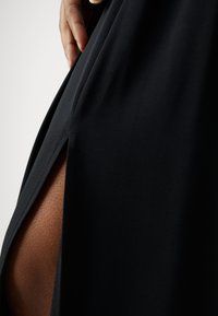 Ruka spočívající na černých šatech z látky s vysokým rozparkem odhalujícím hnědou pleť pod nimi.