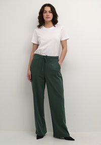 Pantalons larges verts en matériau doux, dotés d'une taille à cordon, de poches latérales et d'une texture lisse. Associés à un t-shirt blanc.