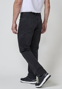 Hombre vestido con jeans negros desgastados y zapatillas negras con suelas blancas, de pie con las piernas cruzadas contra un fondo claro y liso.