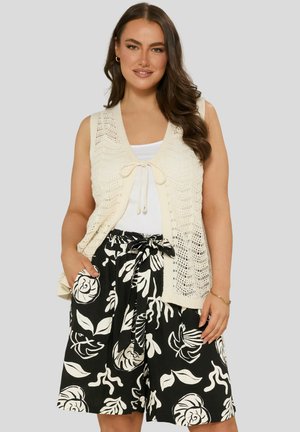 Gilet lavorato a maglia color crema con motivi in pizzo, con una chiusura anteriore a fiocco. Abbinato a pantaloni corti neri con design floreale bianco e una vita annodata.
