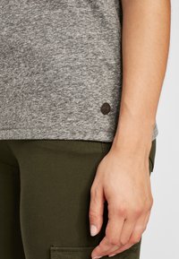 Šedé textured tričko s kulatým výstřihem, krátkými rukávy a malým tmavým logem na lemu. V kombinaci s olivově zelenými upnutými kalhotami.