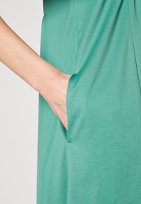 Vêtement vert avec une poche, fabriqué en tissu doux et texturé. Accent sur le détail de la couture de la poche et le tombé fluide du matériau.