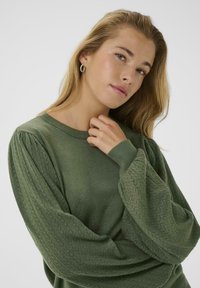 Pull en mailles vert avec des manches bouffantes texturées. Tissu doux, col rond et motifs élaborés sur les poignets.