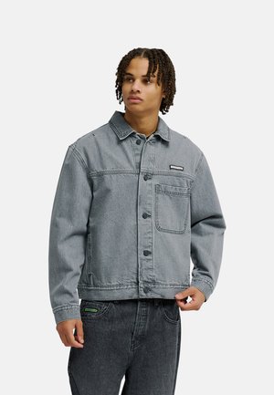 Junger Mann mit kurzen geflochtenen Haaren, trägt eine graue Jeansjacke und dunkelgraue Jeans, blickt leicht zur Seite mit neutralem Ausdruck.