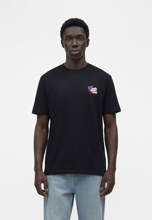 Νέος άνδρας που φοράει μαύρο μπλουζάκι με πολύχρωμο λογότυπο "only and sons" στο στήθος και ανοιχτό μπλε τζιν, στέκεται μπροστά σε έναν απλό λευκό φόντο.