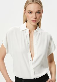 Blusa blanca de manga corta con cuello, cierre delantero de botones y textura suave. Presenta un corte relajado y un escote ligeramente abierto.