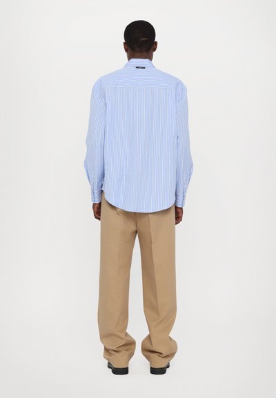 Camisa de rayas azul claro con mangas largas, cuello abotonado y corte relajado; combinada con pantalones beige de pierna ancha y zapatos negros.