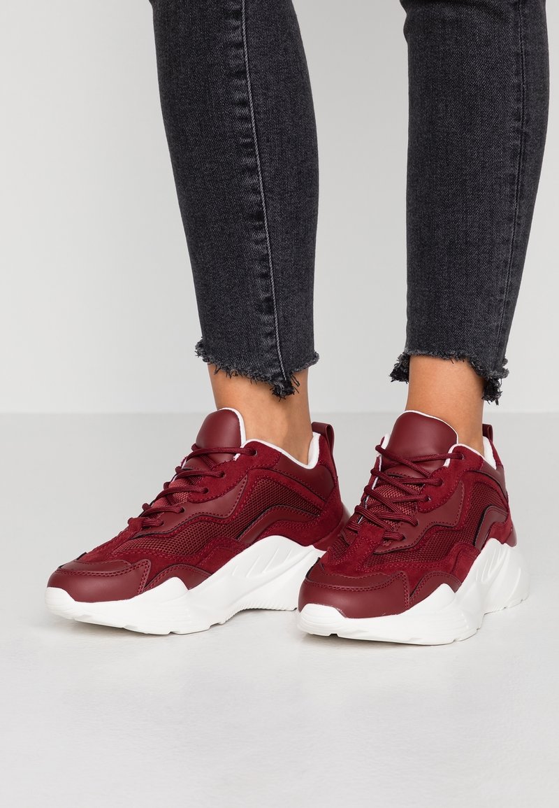 Jambes portant un jean noir à ourlet effiloché et des baskets rouges épaisses avec des semelles blanches épaisses sur un sol gris clair.