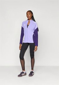 Mulher em pé, vestindo uma jaqueta com fecho éclair roxa clara e roxa escura, leggings de rede pretas e ténis roxos escuros com riscas brancas.