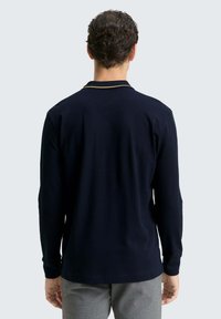 Granatowa koszulka polo z długim rękawem o prążkowanej strukturze, z kontrastującym żółtym wykończeniem kołnierzyka. Gładki tył, połączona z szarymi spodniami.