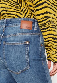 Jeans de mezclilla con un lavado azul, que presentan un parche de cuero marrón etiquetado "DRYKORN", bolsillos laterales y costuras en contraste.