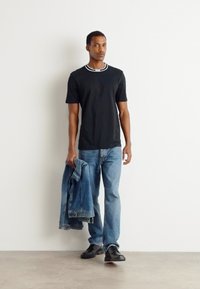 Juodas trumpas rankovėmis marškinėliai su kontrastiniu baltu apykakle, derinami su šviesiai mėlynomis džinsinėmis kelnėmis ir mėlyną džinsinę striukę, laikoma vienoje rankoje.