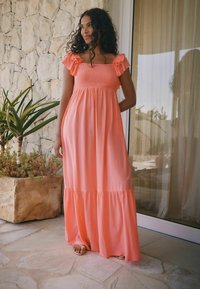 Robe maxi corail avec des bretelles à volants, corsage smocké et jupe à niveaux froncés. Portée avec des sandales plates, devant un mur en pierre.