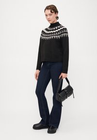 Zwarte gebreide trui met een wit geometrisch patroon, gecombineerd met blauwe flared jeans. Model draagt een kleine zwarte handtas met studs.