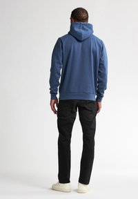 Felpa con cappuccio blu, dal tessuto liscio, con polsini e orlo a costine. Indossata con pantaloni neri e sneakers chiare, vista da dietro.